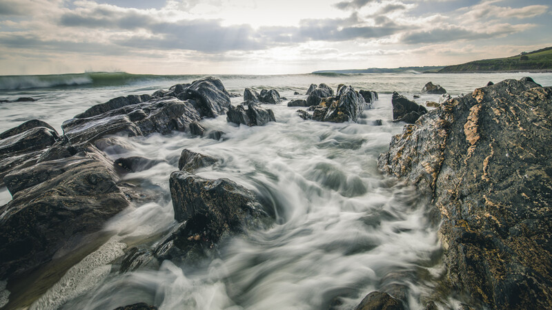 Silky seawater flowing around dark rocky outcrops along a rugged coastline under dramatic clouds.