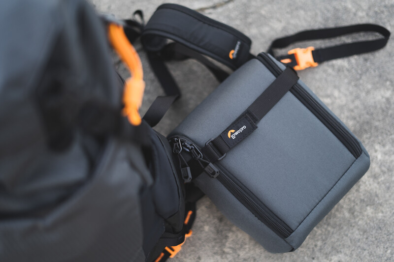 Lowepro camera bags with black and orange accents arranged on pavement next to a person's leg.