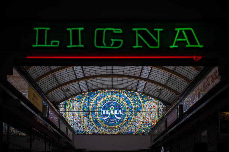 Neon sign reading "LIGNA" above an ornate stained glass window with concentric circular patterns in a industrial ceiling space.
