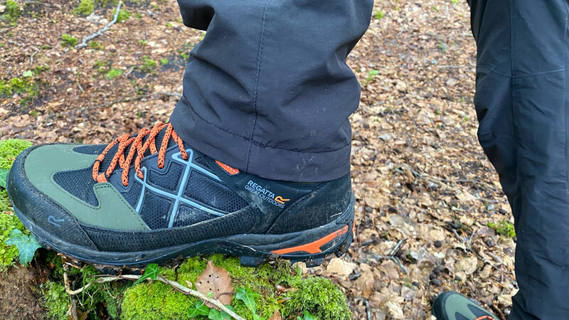 Hiking boot with orange laces on mossy ground next to gravel path.