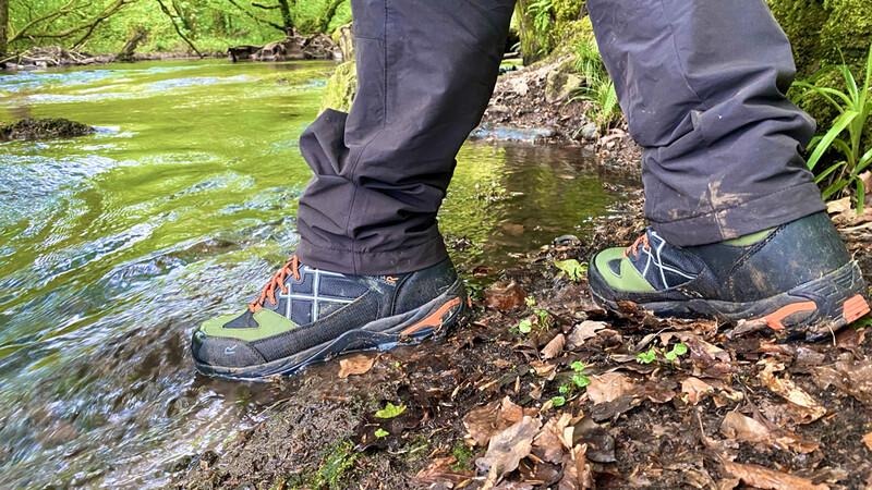Hiker's boots standing on a muddy riverbank beside flowing water with dense green vegetation.