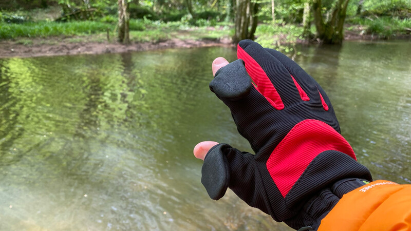 Red and black gloved hand with exposed fingertips pointing toward a calm river surrounded by trees.