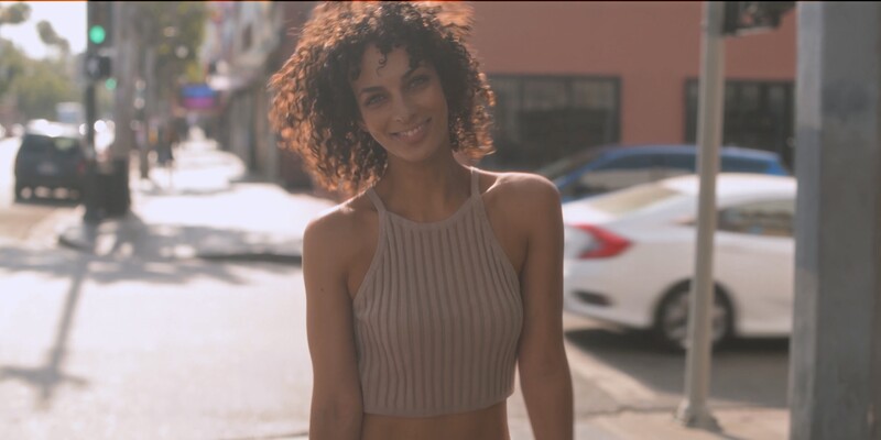 Woman with curly hair smiling at camera on sunny urban street with parked cars.