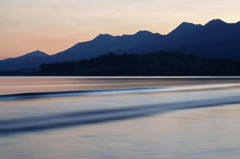 Silhouetted mountains at dusk reflected in calm water with smooth horizontal wave patterns.