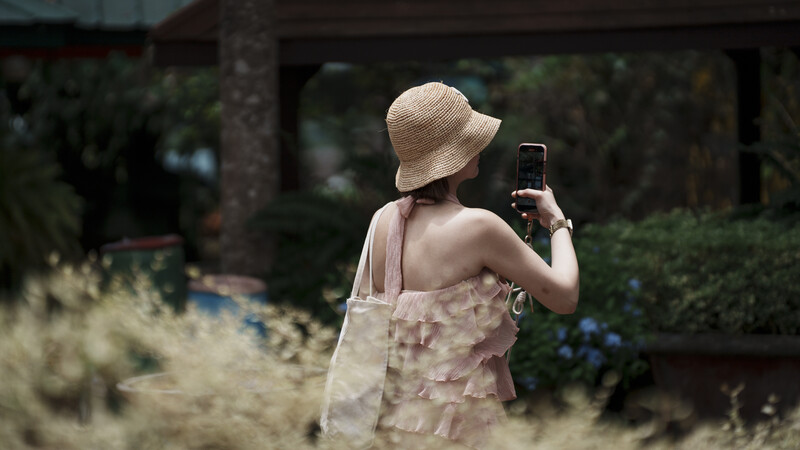 Woman in straw hat holding smartphone while standing in overgrown garden area.