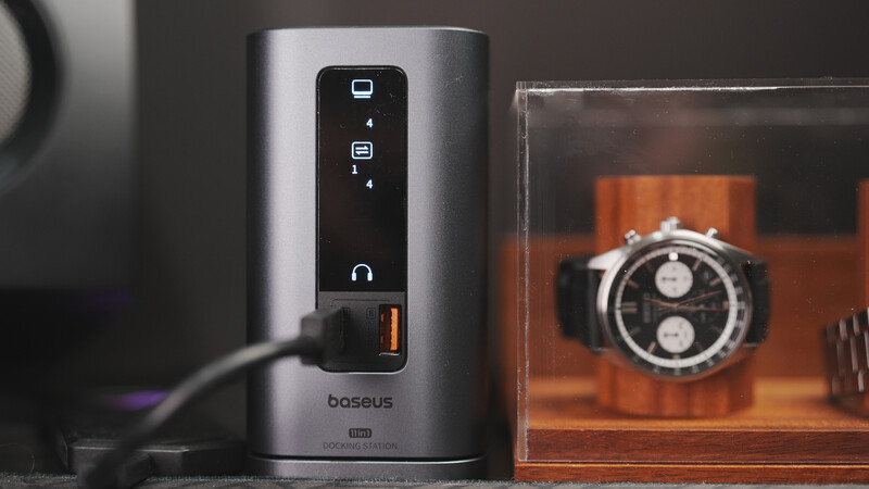 Baseus power strip with multiple USB ports and touchscreen control panel beside a chronograph watch in wooden display case.