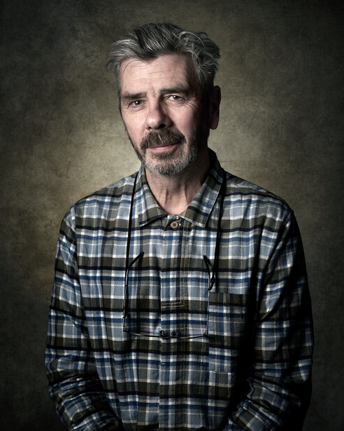 Middle-aged man with gray hair and goatee wearing a blue and white plaid shirt against a muted green background.