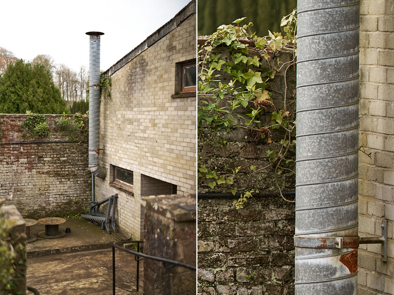 Brick building exterior with metal downpipe and overgrown vegetation alongside ivy-covered wall.