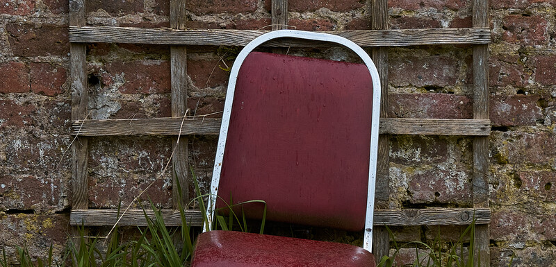 Weathered burgundy chair leaning against a crumbling brick wall with wooden trellis framework.