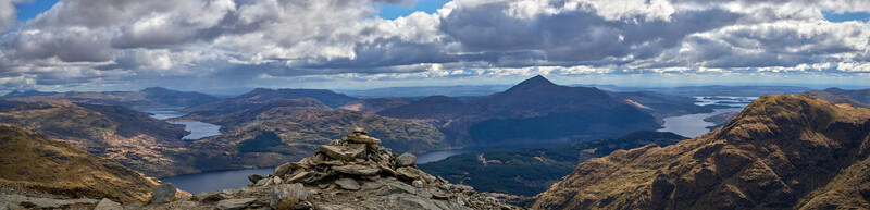 Panoramic mountain landscape with a stone cairn in the foreground overlooking multiple lakes and peaks under partly cloudy skies.