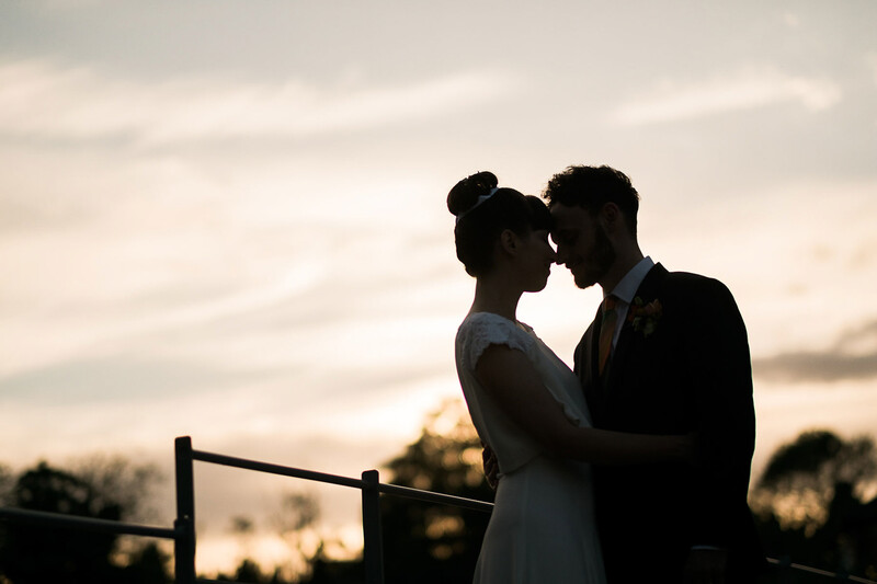 Couple silhouetted against sunset sky, foreheads touching in intimate moment by wooden fence.