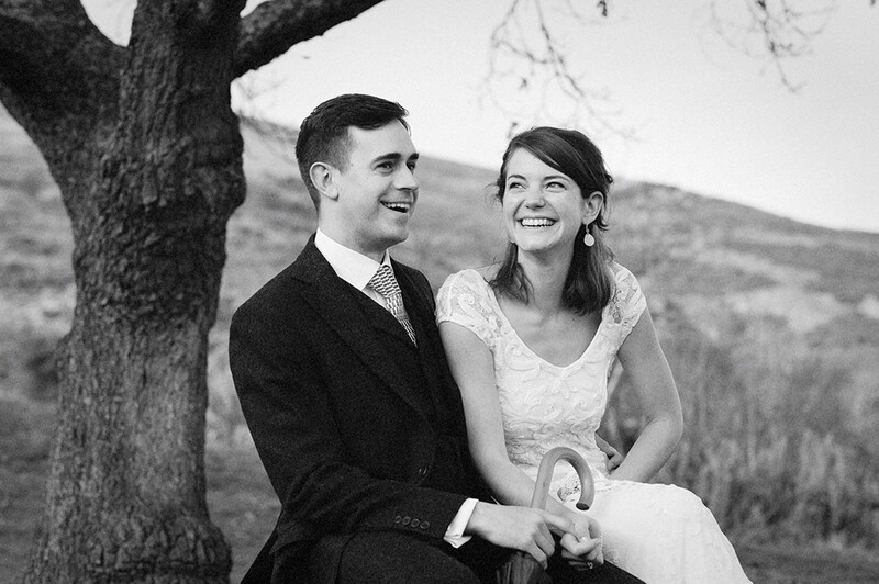 Couple smiling together beneath a large tree in a rural landscape, black and white portrait.