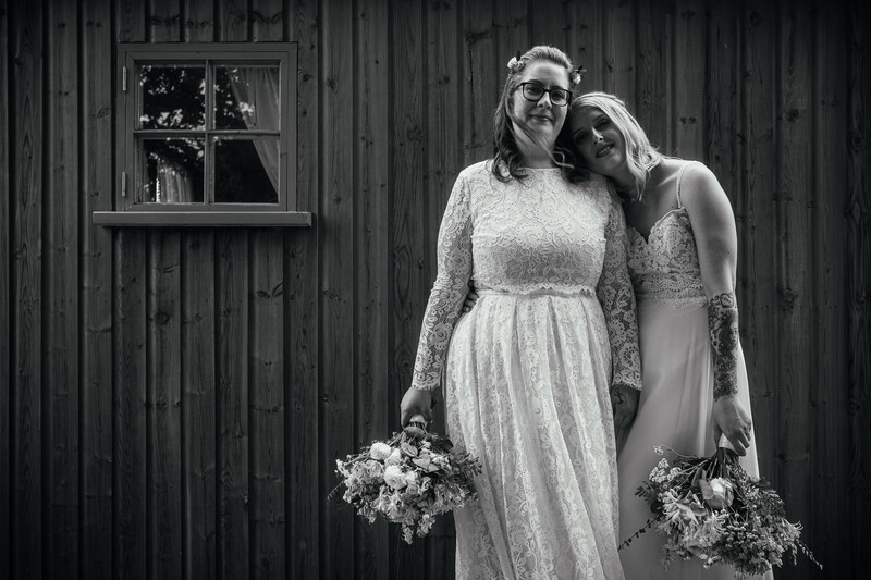 Two brides in white dresses holding bouquets pose against a dark wooden barn wall.