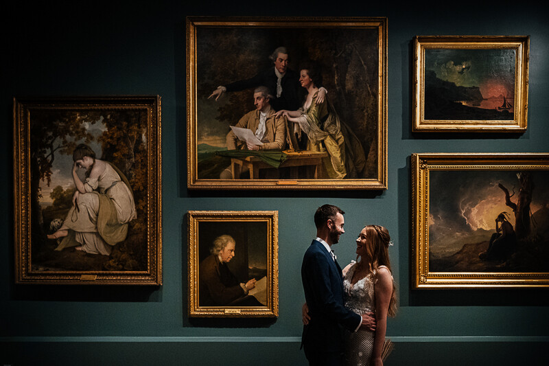 Couple in formal attire viewing classical paintings in a gallery with dark teal walls and gold frames.