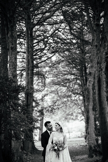 Bride and groom standing together under a tree-lined avenue in black and white.
