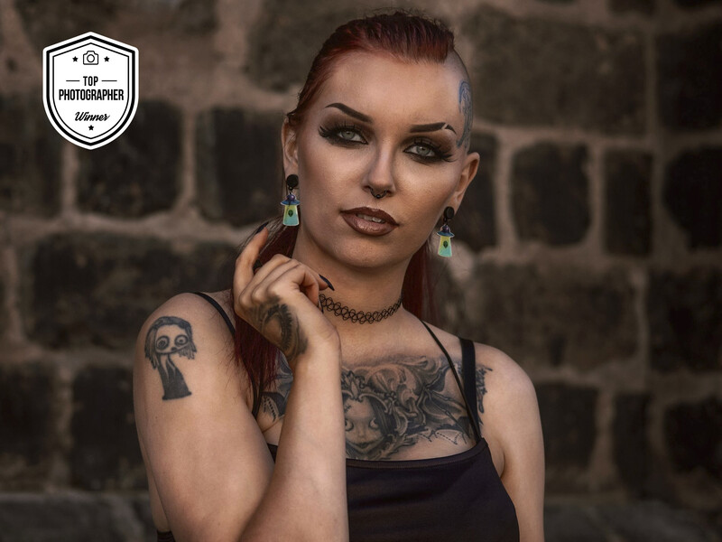 Woman with tattoos posing against a brick wall, wearing dark makeup and chain necklace.