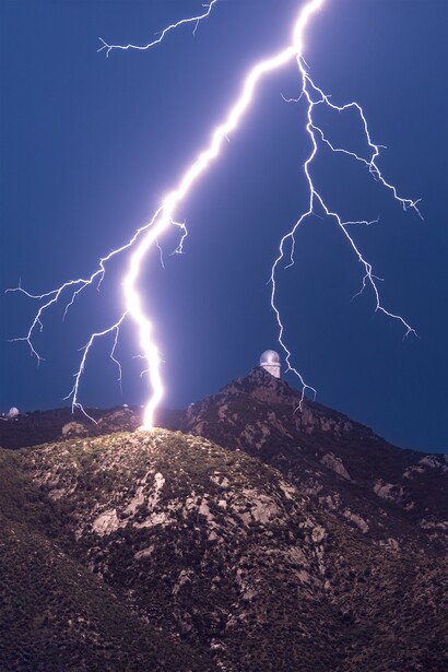 Lightning bolts strike a rocky hilltop against a deep blue night sky.