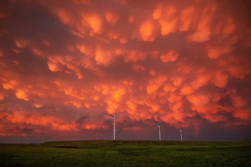 Dramatic orange and red storm clouds dominate the sky above a flat grassland with three distant wind turbines.
