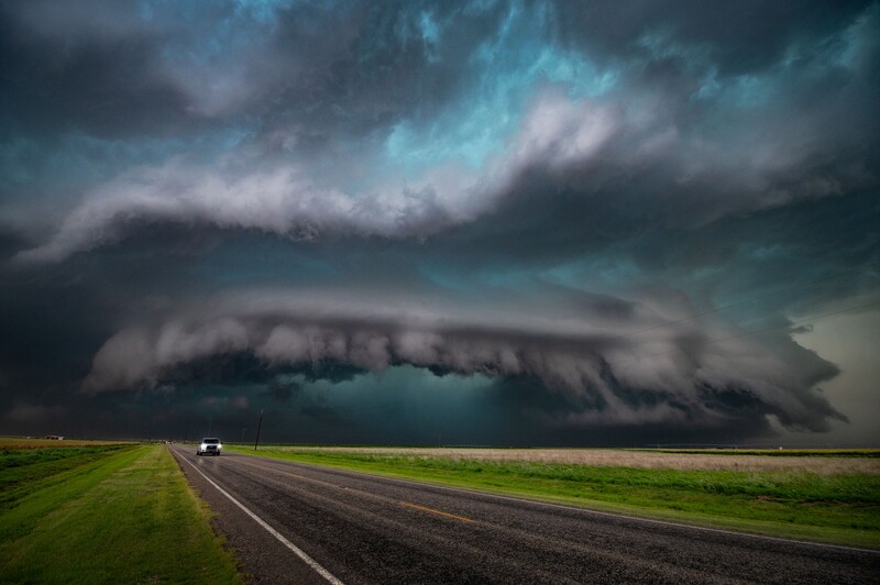 Supercell thunderstorm with dramatic shelf cloud formation looming over a straight highway and green fields.