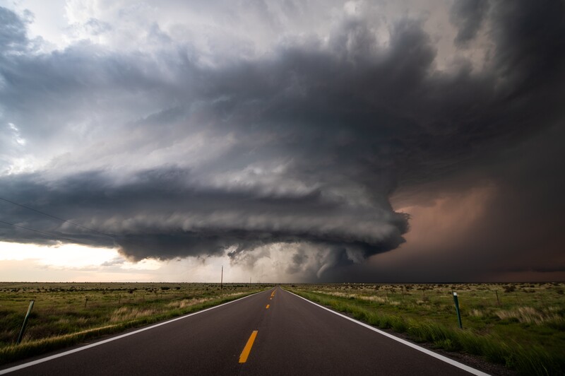 Supercell storm with anvil clouds looming over a straight highway stretching across flat plains.