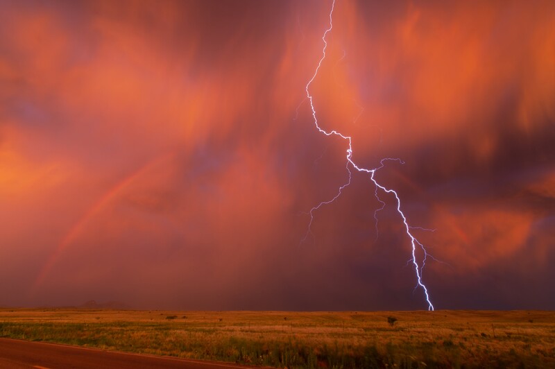 Lightning bolt striking across an orange and red storm sky above a flat grassland at dusk.