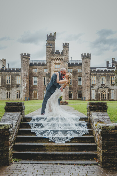 Bride in wedding dress with flowing veil posing on stone steps in front of castle.