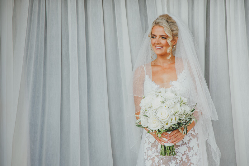 Bride in lace wedding dress and veil holding white floral bouquet against sheer curtain backdrop.