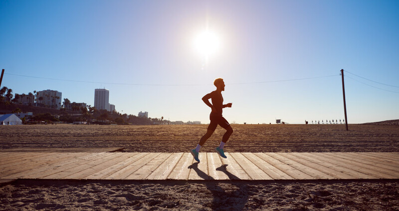 Runner jogging on a wooden boardwalk at a beach during bright daylight with cityscape in background.