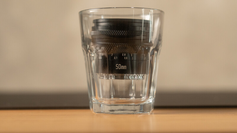 A 50mm camera lens standing upright inside a clear drinking glass on a wooden surface.