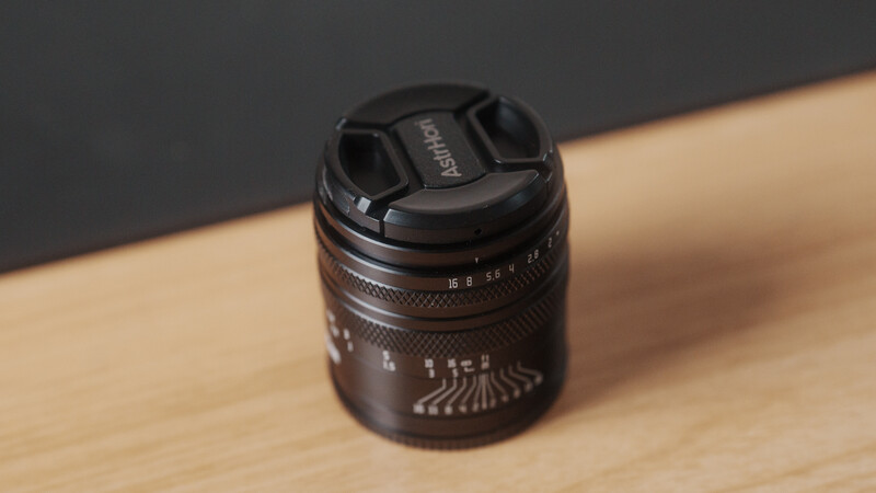 Dark lens with focus ring and aperture markings sitting on light wood surface against gray background.
