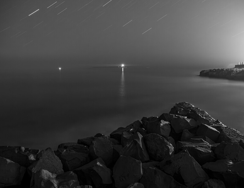 Dark seascape with rocky breakwater in foreground, distant lighthouse beams, and star trails across night sky.