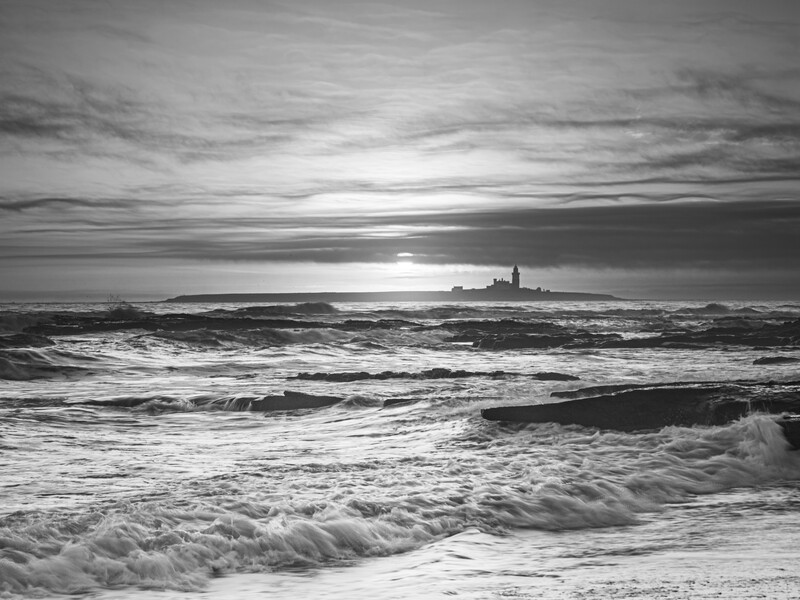 Monochrome seascape with waves rolling onto a sandy beach under a dramatic cloudy sky at sunset.