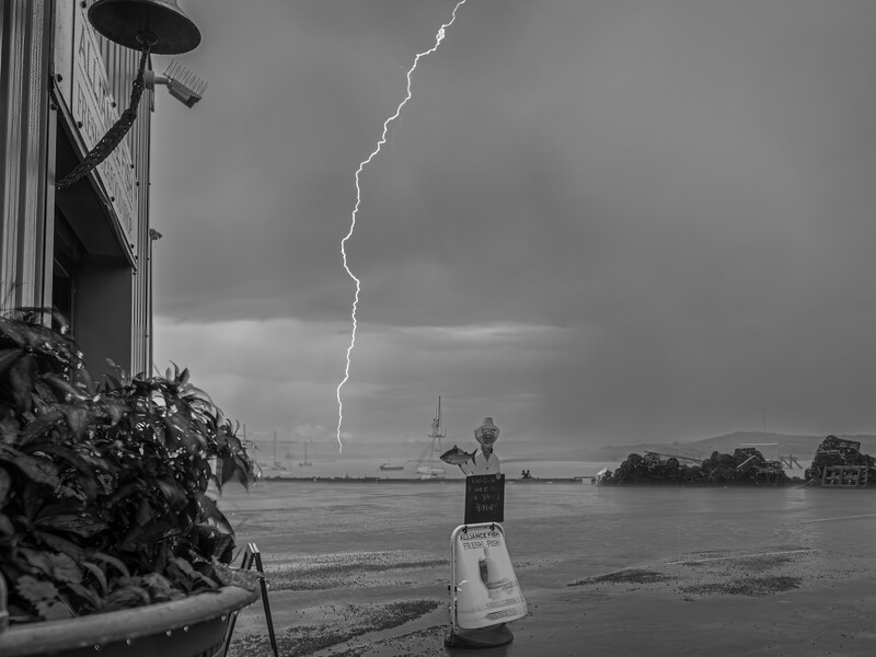 Lightning bolt strikes across stormy sky above a waterfront with moored boats and industrial structures.