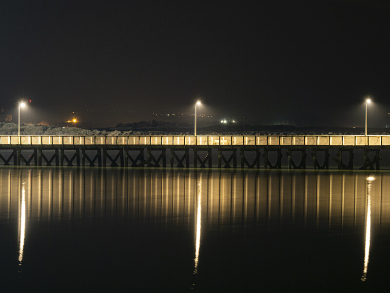 Illuminated bridge structure reflected in still dark water at night.
