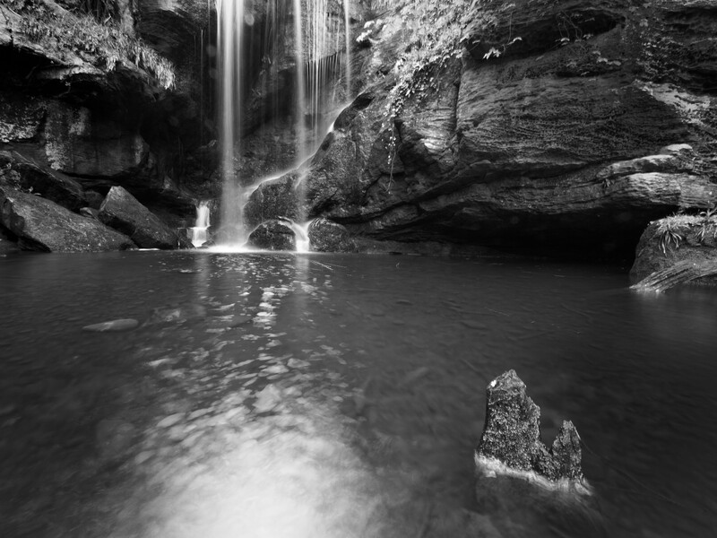 Waterfall cascading into a dark pool surrounded by layered rock formations in black and white.