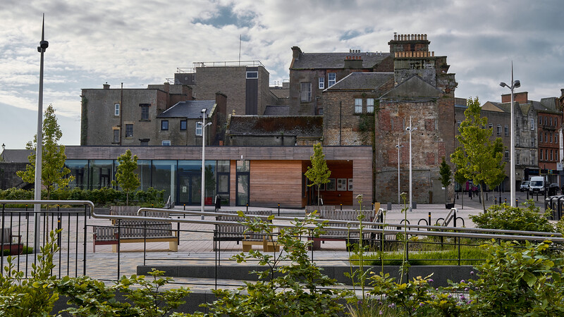 Waterfront plaza with modern pavilion building backed by historic stone architecture under cloudy sky.