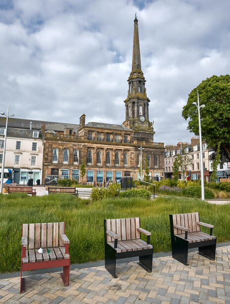 Three wooden Adirondack chairs on a paved plaza facing a sandstone church with a tall spire.