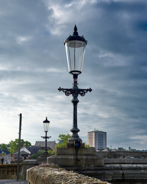 Ornate black street lamp on stone pedestal beside river with cloudy sky and distant buildings.
