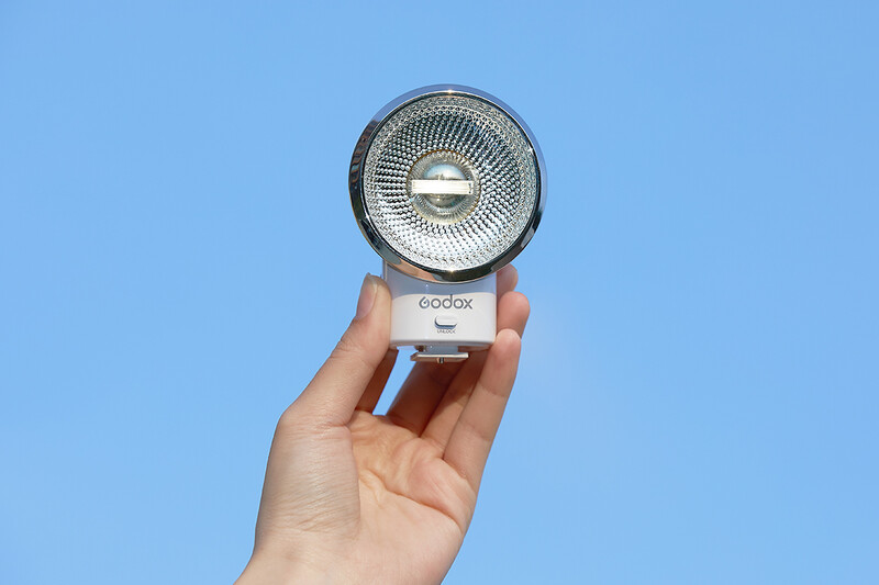 Hand holding a compact Godox camera flash against a clear blue sky background.