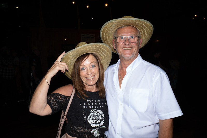 Two people wearing straw hats pose together at an evening event against a dark background.