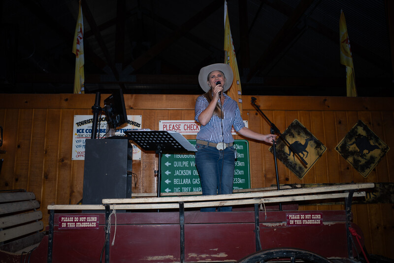 Singer in cowboy hat performing at wooden venue stage with microphone and visible scoreboards.