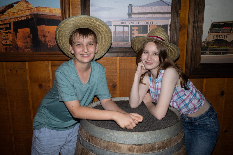 Two young children wearing straw cowboy hats pose beside a wooden barrel in a rustic interior setting.
