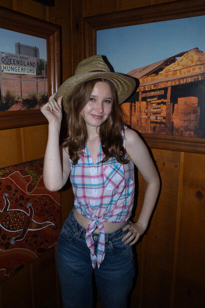 Woman in plaid shirt and cowboy hat posing indoors with rustic wooden interior and mountain landscape visible through windows.