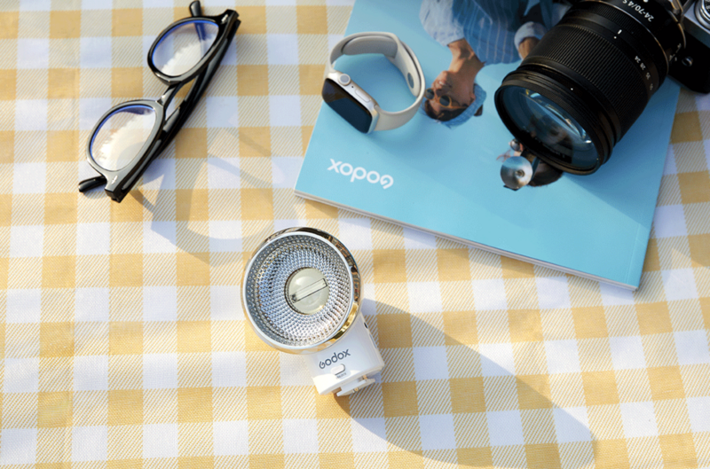 Flat lay of photography gear on a checkered yellow and white fabric background featuring eyeglasses, a camera lens, a smartwatch, and a blue product box.