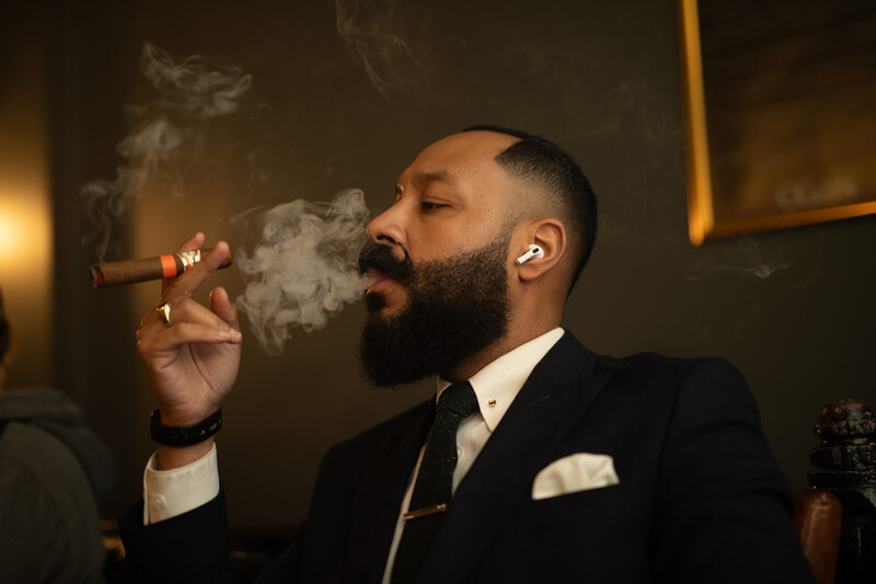 Bearded man in black suit and tie smoking cigar in warm interior lighting.