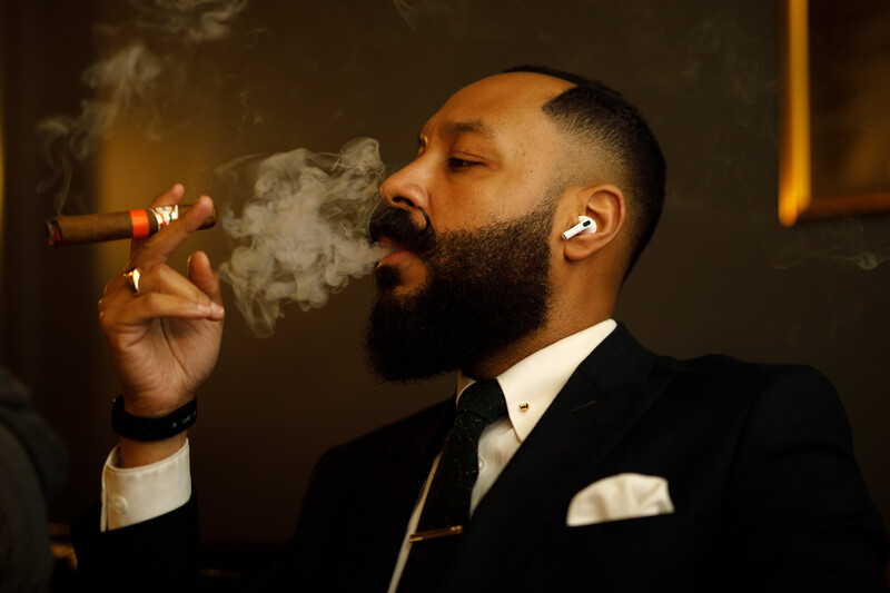 Man in formal attire smoking a cigar with dramatic warm lighting against a dark background.