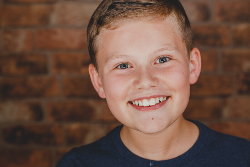 Young boy with light brown hair smiling at camera against brick wall backdrop.