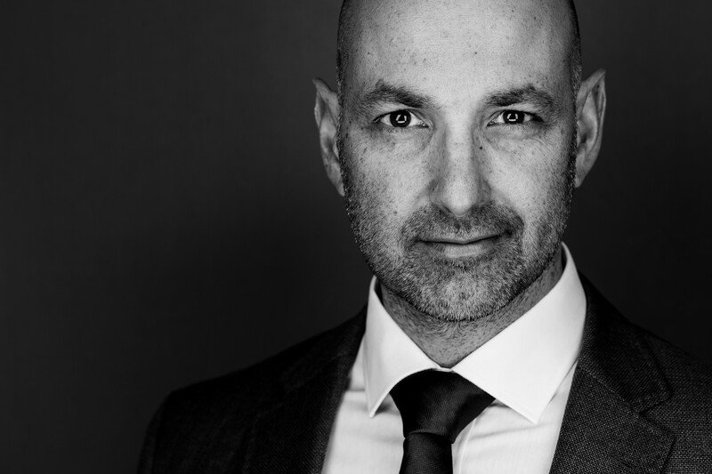 Professional headshot of a bald man with facial hair wearing a dark suit and black tie against a dark background.