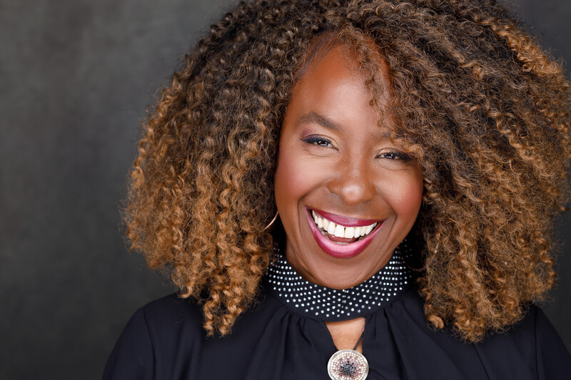 Woman with voluminous curly brown hair smiling at camera against dark background.