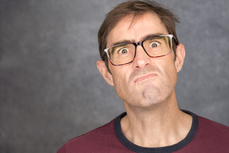 Man wearing glasses and maroon shirt with skeptical expression against gray background.